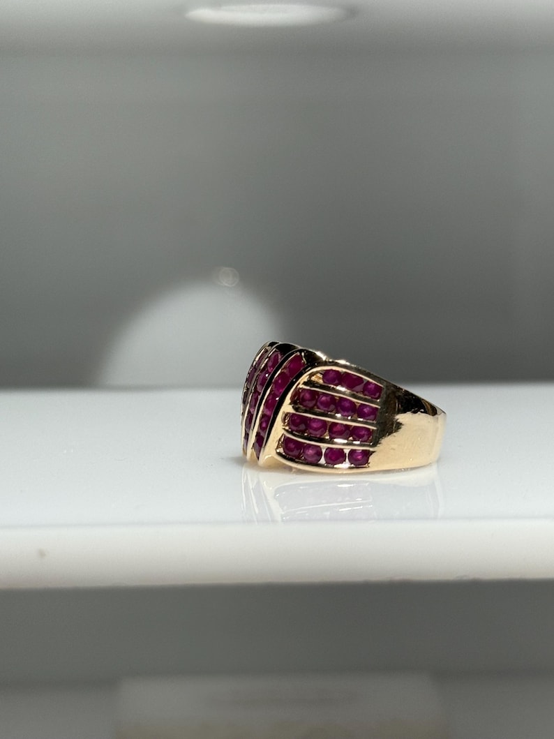 K&ouml;nnte beinhalten: Ein goldener Ring mit einer Ansammlung kleiner, runder, rubinroter Steine. Die Steine sind in Reihen angeordnet und bilden eine strukturierte Oberfl&auml;che. Der Ring hat einen dicken Schaft und ein poliertes Finish.