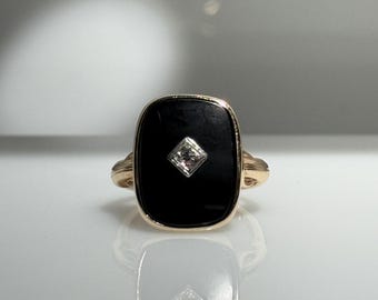 May include: A gold ring with a rectangular black onyx stone and a square diamond at its centre. The ring has a classic design with a polished finish, highlighting the gemstone's brilliance.