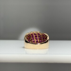 K&ouml;nnte beinhalten: Ein goldener Ring mit einem Band aus kleinen, quadratisch geschliffenen Rubinen. Der Ring hat ein dickes, poliertes Band mit glatter, reflektierender Oberfl&auml;che. Die Rubine sind tiefrot und eng aneinander gesetzt, um eine durchgehende Oberfl&auml;che zu bilden.