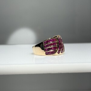 K&ouml;nnte beinhalten: Ein goldener Ring mit einer Ansammlung kleiner, leuchtend roter Edelsteine. Das Design des Rings weist mehrere Reihen der roten Steine auf, wodurch ein strukturiertes und auff&auml;lliges Aussehen entsteht. Das Band ist goldfarben poliert.