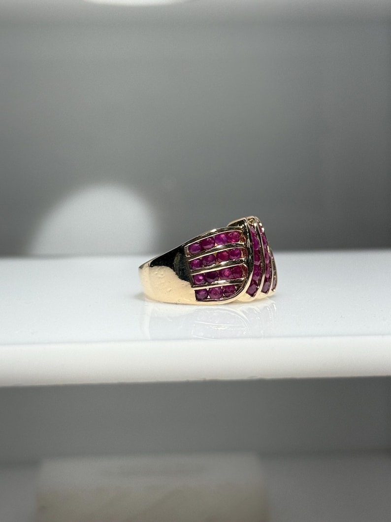 K&ouml;nnte beinhalten: Ein goldener Ring mit einer Ansammlung kleiner, leuchtend roter Edelsteine. Das Design des Rings weist mehrere Reihen der roten Steine auf, wodurch ein strukturiertes und auff&auml;lliges Aussehen entsteht. Das Band ist goldfarben poliert.