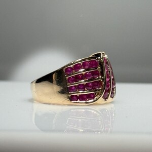 K&ouml;nnte beinhalten: Ein goldener Ring mit mehreren Reihen kleiner, runder, leuchtend roter Edelsteine. Der Ring hat ein breites Band und eine gew&ouml;lbte Oberseite, wobei die Edelsteine eng beieinander liegen. Der Ring wird auf einer reflektierenden Oberfl&auml;che pr&auml;sentiert.