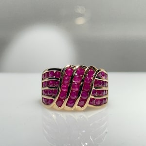 K&ouml;nnte beinhalten: Ein goldener Ring mit mehreren Reihen leuchtend roter Rubine. Die Rubine sind in einem wellenartigen Muster angeordnet, wodurch ein strukturiertes und auff&auml;lliges Design entsteht. Der Ring wird auf einer reflektierenden Oberfl&auml;che gezeigt.