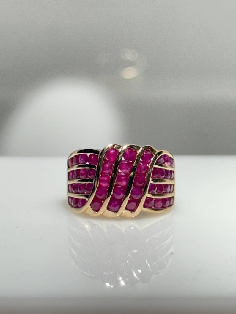 K&ouml;nnte beinhalten: Ein goldener Ring mit mehreren Reihen leuchtend roter Rubine. Die Rubine sind in einem wellenartigen Muster angeordnet, wodurch ein strukturiertes und auff&auml;lliges Design entsteht. Der Ring wird auf einer reflektierenden Oberfl&auml;che gezeigt.
