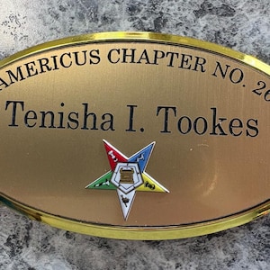 May include: An oval-shaped gold-toned name badge with a polished gold border. The text reads AMERICUS CHAPTER NO. 26 and Tenisha I. Tookes in black lettering. A multi-coloured five-pointed star emblem with a central tower icon is centred below.