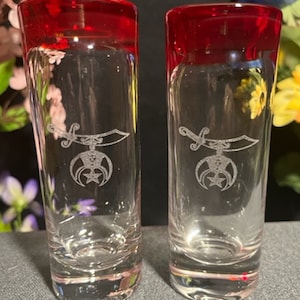 May include: Two clear glass shot glasses with red rims. Each glass has an etched design of a sword and a crescent moon with a star.