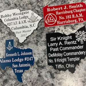 May include: Four state-shaped plaques featuring custom text and Masonic emblems. One white South Carolina, one red Pennsylvania, one blue Texas, and one black Ohio plaque, each engraved with specific names, lodge titles, and locations in white text.