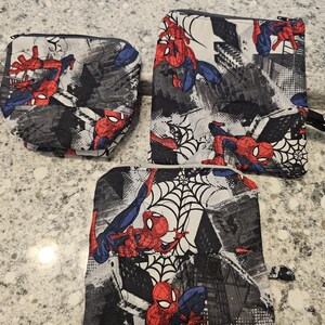 May include: Three zippered pouches featuring a Spider-Man comic book design. The pouches have a black, gray, and white background with Spider-Man in red and blue. The pouches are different sizes and shapes.