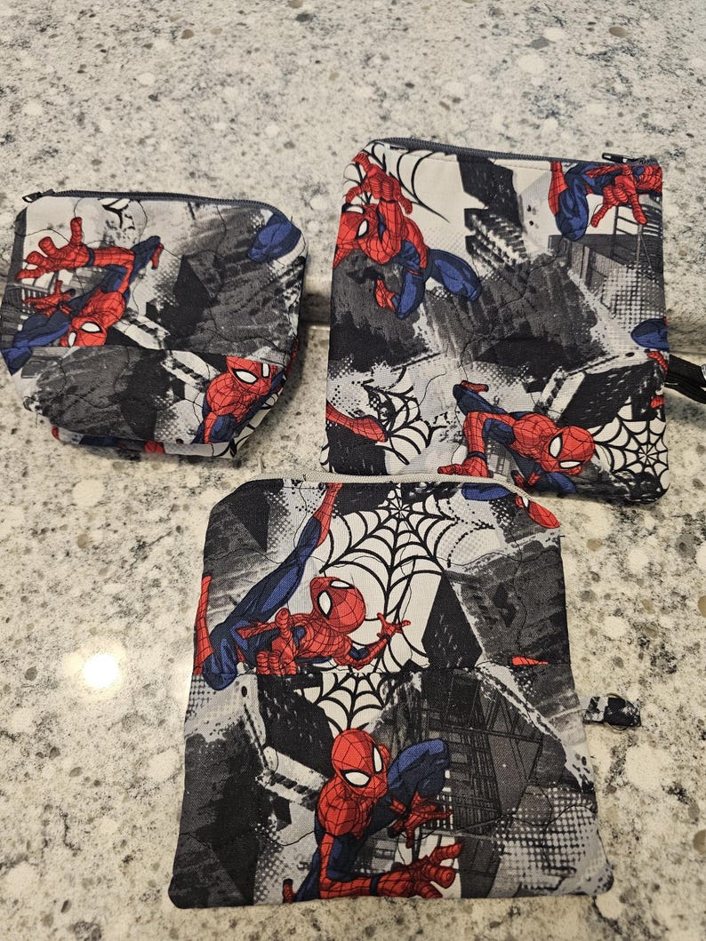May include: Three zippered pouches featuring a Spider-Man comic book design. The pouches have a black, gray, and white background with Spider-Man in red and blue. The pouches are different sizes and shapes.