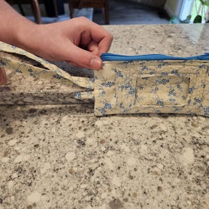 May include: A small, quilted pouch with a blue zipper and a matching wrist strap. The pouch is cream-colored and decorated with a delicate blue floral pattern. The wrist strap is attached to the side of the pouch.