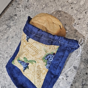 May include: A round loaf of bread sits inside a blue and beige bread bag. The bag features a grape design and text. The bag is made of fabric and has a drawstring closure. The bread is golden brown and has a crusty appearance.