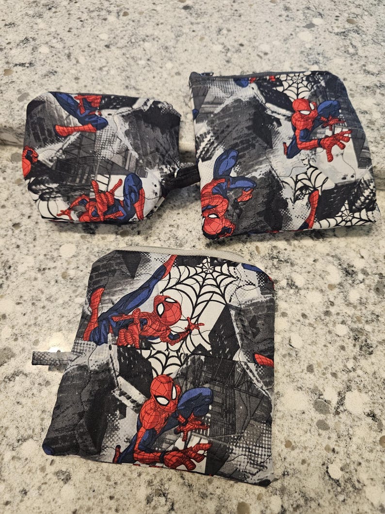 May include: Three Spider-Man themed pouches. The pouches feature a repeating pattern of Spider-Man in action poses, along with spiderwebs and cityscapes. The color scheme is primarily red, blue, black, and white.