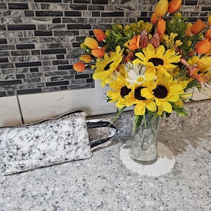 May include: A floral print cosmetic bag with a black handle sits next to a vase of sunflowers, orange tulips, and other flowers. The bag is white with a black floral pattern. The flowers are in a clear glass vase.