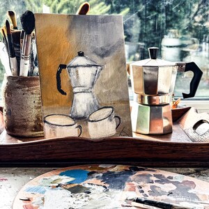 May include: A still life featuring a painting of a coffee pot and cups, a metal stovetop espresso maker, and a wooden artist's palette with paint. Paintbrushes are in a ceramic jar. The scene is set near a window with a view of trees.
