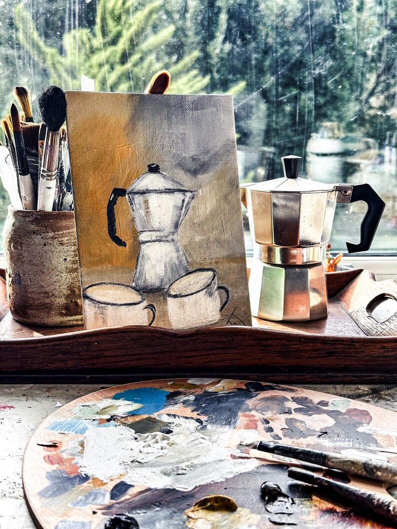 May include: A still life featuring a painting of a coffee pot and cups, a metal stovetop espresso maker, and a wooden artist's palette with paint. Paintbrushes are in a ceramic jar. The scene is set near a window with a view of trees.