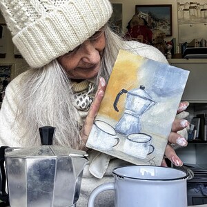 May include: A silver stovetop espresso maker, a white enamel mug with a black rim, and a small painting of a coffee pot and cups. The painting has a yellow and blue background. A person wearing a white knit hat is holding the painting.