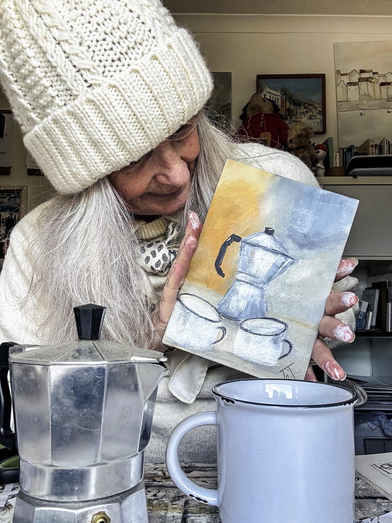 May include: A silver stovetop espresso maker, a white enamel mug with a black rim, and a small painting of a coffee pot and cups. The painting has a yellow and blue background. A person wearing a white knit hat is holding the painting.