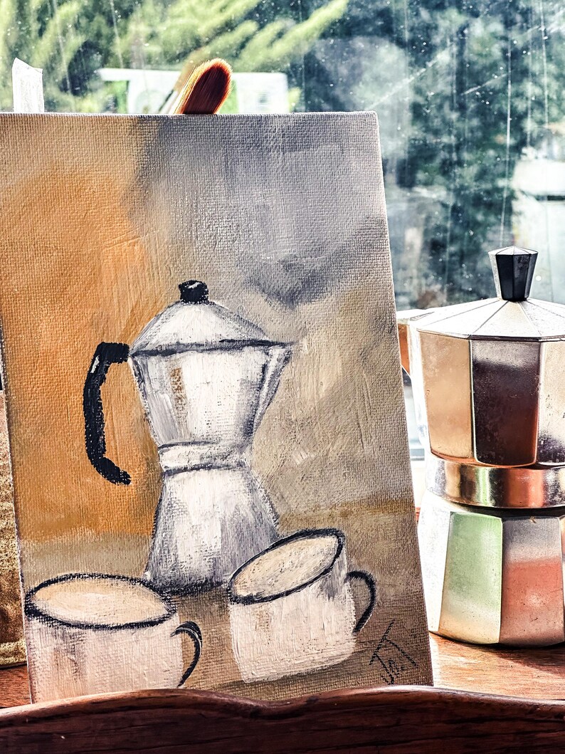 May include: An oil painting on canvas shows a coffee pot and two mugs in neutral tones. A metal stovetop espresso maker sits beside the painting. A paintbrush rests on top of the canvas.