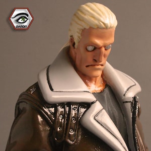 May include: A close-up of a sculpted figure with a white and brown leather jacket. The figure has short, blonde hair and a serious expression. The figure's eyes are painted with a light blue color.