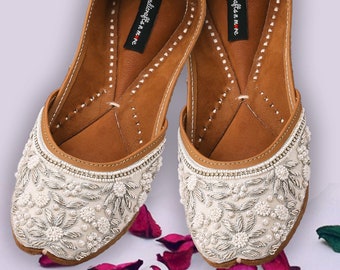 May include: A pair of white embroidered flat shoes with silver embellishments and pearl accents. The shoes have a brown leather insole with the text "Handicrafts n me" printed on them.