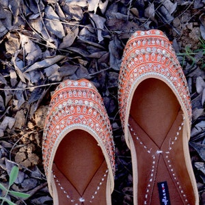 May include: A pair of orange and red embroidered flat shoes with a pointed toe. The shoes have a decorative design with silver sequins and a red trim. The brand name "handicrafts" is printed on the inside of the shoes.