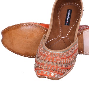 May include: A pair of orange and gold embroidered flat shoes with a leather sole. The shoes have a pointed toe and are decorated with sequins and beads. The brand name "Handicrafts & More" is visible on the inside of the shoe.