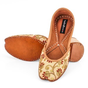May include: A pair of beige embroidered flat shoes with red and gold accents. The shoes have a leather sole and are decorated with floral patterns. The brand name "Handicrafts n me" is printed on the inside of one shoe.