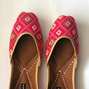May include: A pair of pink and green flat shoes with a geometric pattern. The shoes have a gold trim and a brown leather insole. The brand name "handicrafts n more" is printed on the insole.