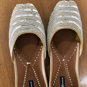 May include: A pair of gold and silver sequined flat shoes with a brown leather insole. The shoes have a pointed toe and a small strap across the top. The insole is labeled "handicrafts in me".