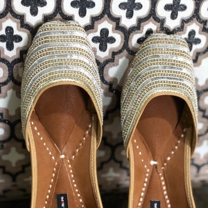 May include: Pair of gold and silver embellished flat shoes with a pointed toe and a brown leather insole. The shoes have a decorative stitching detail around the edge of the insole. The insole is labeled "handicrafts n more".