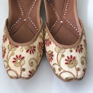 May include: A pair of cream-colored embroidered flat shoes with red and gold floral designs. The shoes have a brown leather insole and a pointed toe.