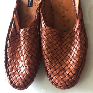 May include: A pair of brown leather woven slippers with a black label that reads "Handicrafts n more".