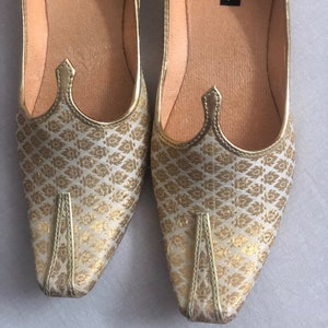 May include: A pair of gold and white patterned flat shoes with pointed toes. The shoes have a decorative detail on the top and the words "handicrafts in me" are printed on the inside of the shoes.