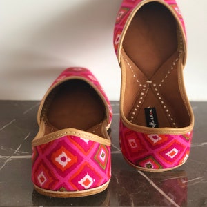 May include: A pair of pink and green patterned flat shoes with a pointed toe and a decorative stitch detail. The shoes have a brown leather insole and a gold trim.