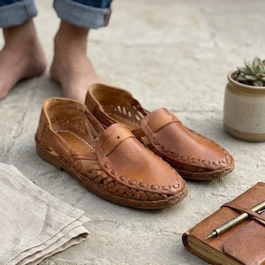 May include: A pair of tan leather huarache-style slip-on shoes featuring a woven lattice design across the upper, a rounded toe, and a stitched leather strap detail across the vamp. The shoes have a low-profile construction with visible stitching.
