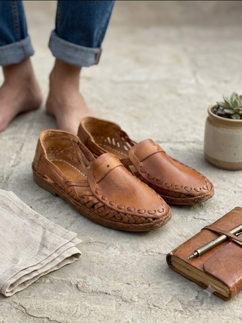 May include: A pair of tan leather huarache-style slip-on shoes featuring a woven lattice design across the upper, a rounded toe, and a stitched leather strap detail across the vamp. The shoes have a low-profile construction with visible stitching.
