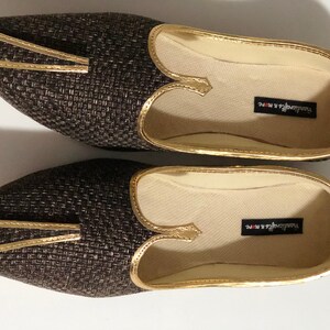 May include: A pair of brown woven flat shoes with gold trim and a pointed toe. The shoes have a cream-colored interior and the text "Trendsetter & More" is printed on the inside of each shoe.