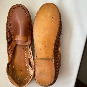 May include: A pair of brown leather huarache sandals. One sandal is positioned to show the smooth leather upper with woven side detailing and a strap across the vamp, while the other is flipped to display the flat, tan leather sole and stitching.