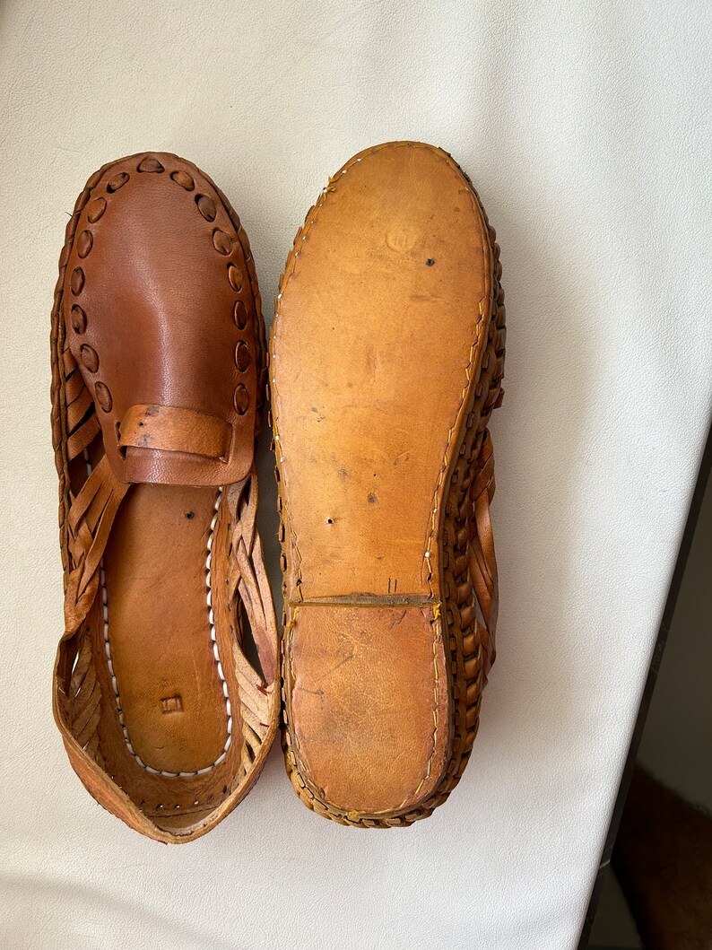 May include: A pair of brown leather huarache sandals. One sandal is positioned to show the smooth leather upper with woven side detailing and a strap across the vamp, while the other is flipped to display the flat, tan leather sole and stitching.