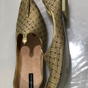 May include: A pair of gold woven flat shoes with a pointed toe and a small decorative piece on the top. The shoes have a label that reads "handicrafts n' me".