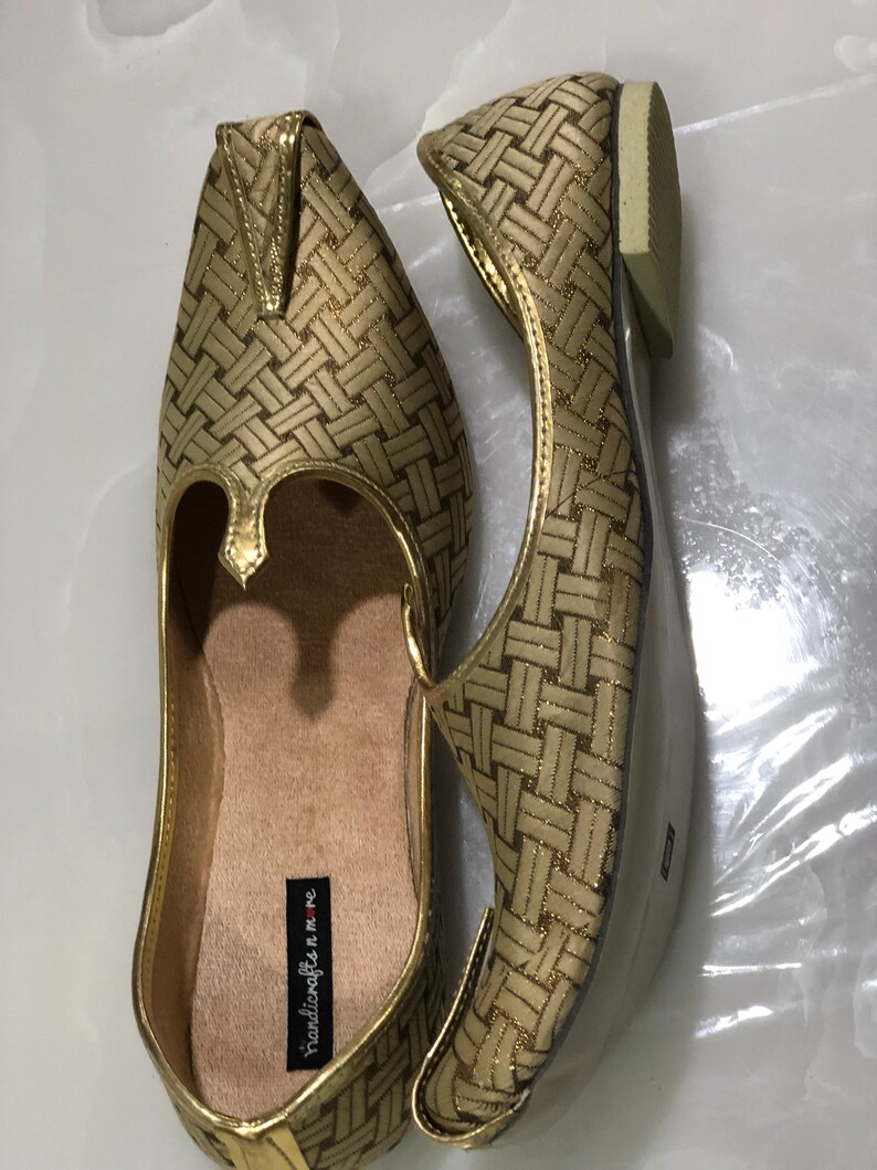 May include: A pair of gold woven flat shoes with a pointed toe and a small decorative piece on the top. The shoes have a label that reads "handicrafts n' me".