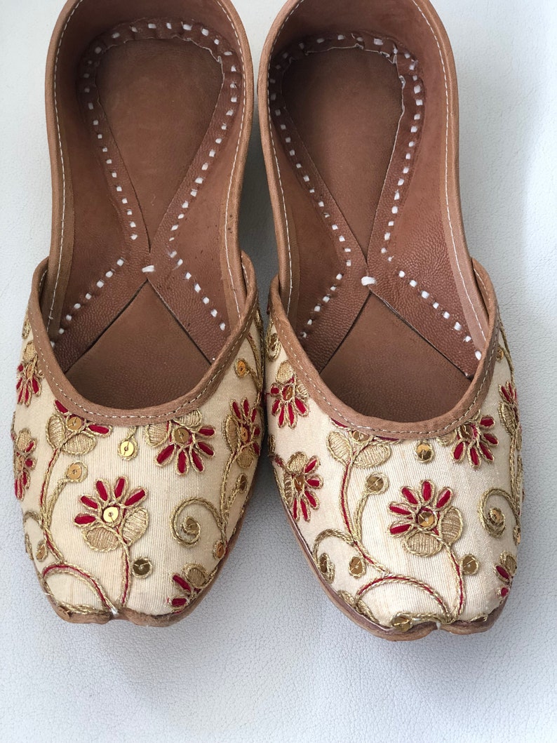 May include: A pair of cream-colored embroidered flat shoes with red and gold floral designs. The shoes have a brown leather insole and a pointed toe.