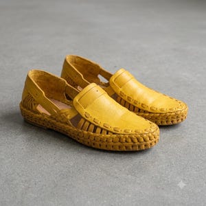 May include: A pair of mustard yellow leather loafers featuring a woven design along the sides and base. The shoes have a closed toe, an open-sided construction with cutouts, and a thick, braided leather trim that encircles the entire outer edge.
