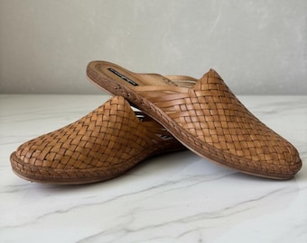 May include: A pair of tan leather mules featuring a woven upper design with a closed, rounded toe and a flat sole. The interior footbed is lined with matching tan leather and includes a visible brand label near the heel area of the shoe.