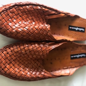 May include: A pair of brown leather woven slippers. The slippers have a woven leather upper and a leather sole. The brand name "Sandals" is visible on the sole.