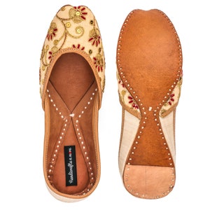May include: A pair of beige leather flats with red and gold embroidery. The shoes have a pointed toe and a low heel. The brand name "handicrafts n more" is printed on the insole.