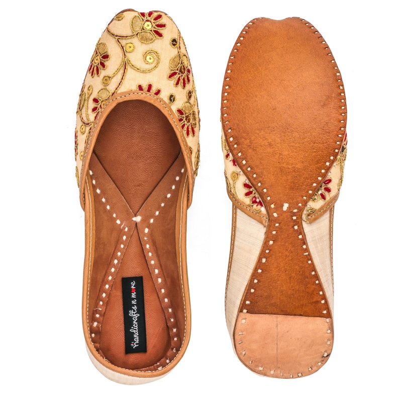 May include: A pair of beige leather flats with red and gold embroidery. The shoes have a pointed toe and a low heel. The brand name "handicrafts n more" is printed on the insole.