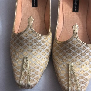 May include: A pair of gold and white patterned flat shoes with a pointed toe. The shoes have a gold trim and the words "handicrafts in me" are printed on the inside of each shoe.