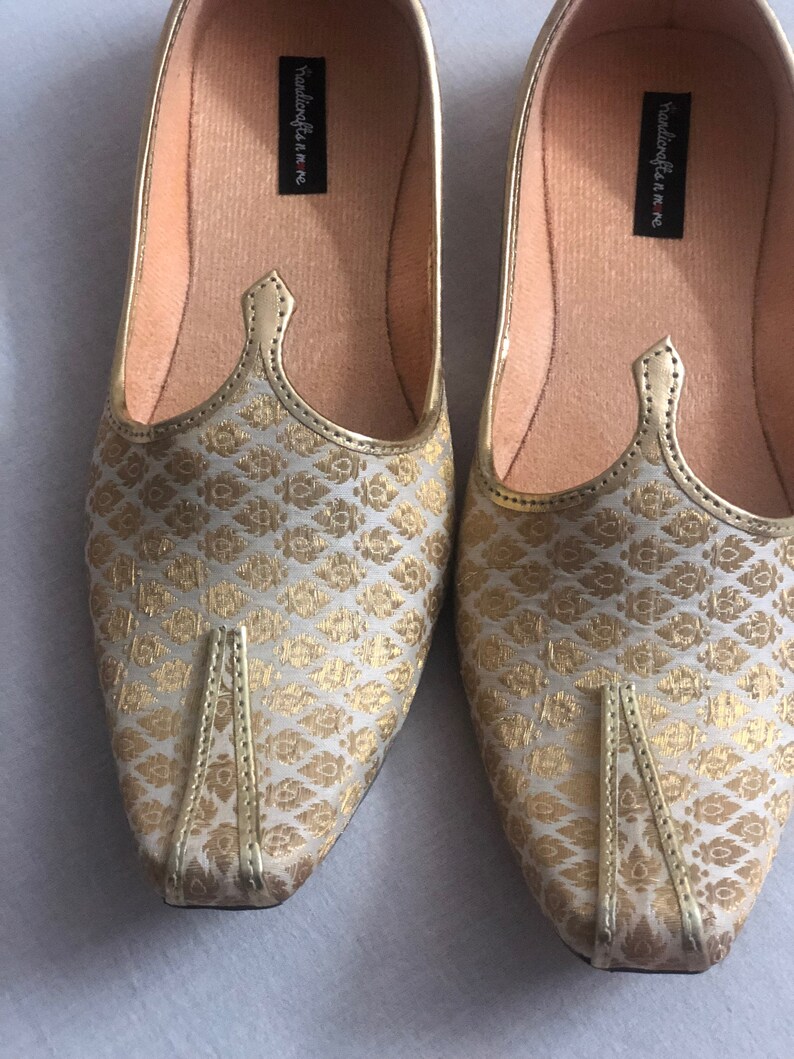 May include: A pair of gold and white patterned flat shoes with a pointed toe. The shoes have a gold trim and the words "handicrafts in me" are printed on the inside of each shoe.