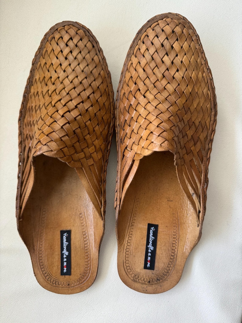 May include: A pair of light brown leather slip-on shoes. The shoes feature a woven design across the top and sides. The soles are a lighter shade of brown. The shoes have a label that reads "Handicrafts n m e".